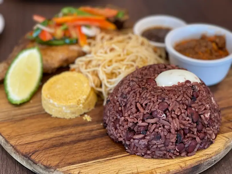 Üzerinde haşlanmış yumurta olan kubbe şeklinde kırmızımsı kahverengi pirinç ve börülce; yanında spagetti, kızarmış balık, gari, misket limonu ve iki sos bulunan bir servis tahtası