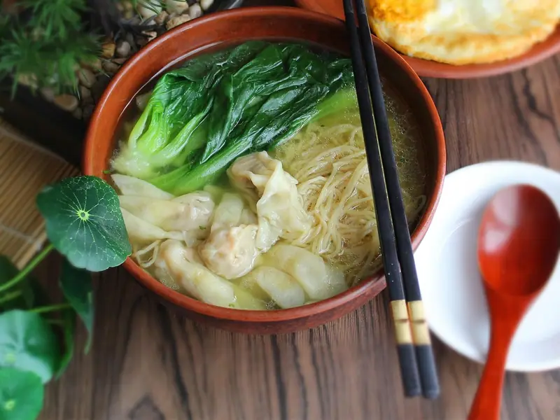 Schale Wan-Tan-Nudelsuppe mit Pak Choi und dünnen Eiernudeln in klarer Brühe, daneben Essstäbchen, ein Spiegelei und ein Schälchen Sojasauce
