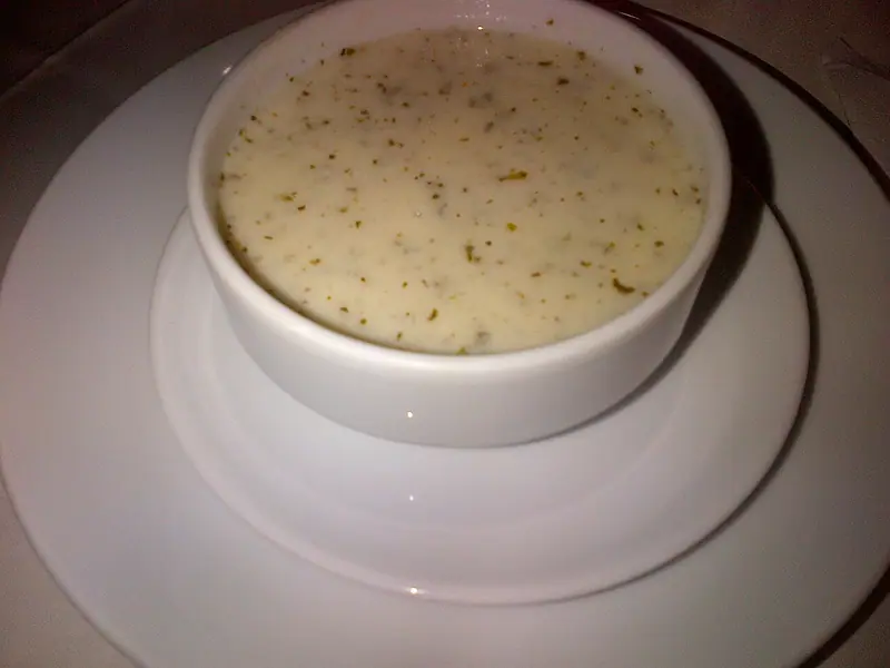 A creamy pale soup speckled with dried herbs in a white ramekin-style bowl set on a white saucer and plate.