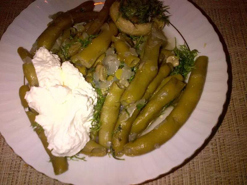 Braised green bean pods with onion and fresh dill on a white plate, topped with a dollop of thick white yogurt on one side.