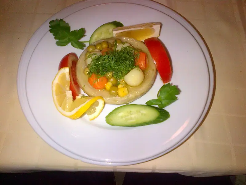 Bonggol artichoke berisi kacang polong, wortel, kentang, dan jagung, ditaburi daun dill, dengan lemon, tomat, mentimun, dan peterseli di piring putih.