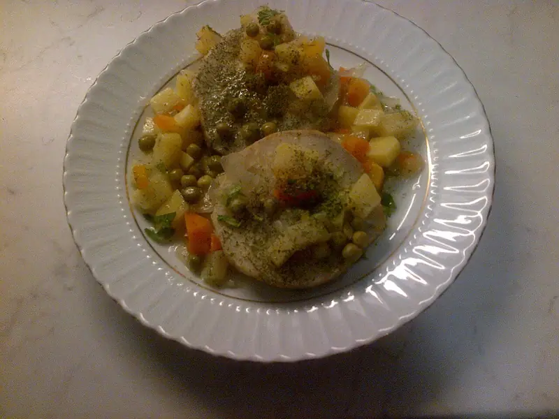 Two thick celeriac slices topped with dried dill, surrounded by diced potato or celeriac, carrot and peas on a white scalloped plate.