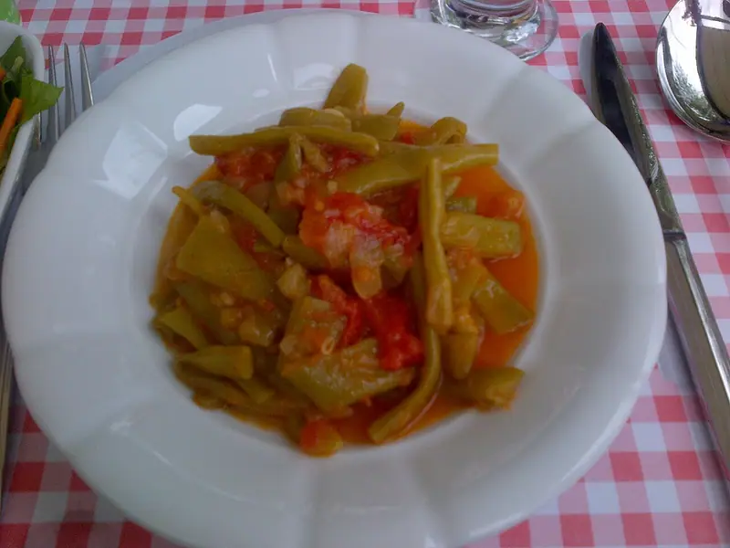 Vagem-manteiga refogada em um molho leve de tomate e cebola, servida em uma tigela branca de borda ondulada sobre uma toalha xadrez vermelha, com talheres ao lado.